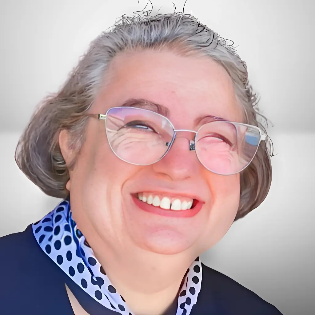 Person with gray hair wearing glasses and a blue polka dot blouse under a blazer.